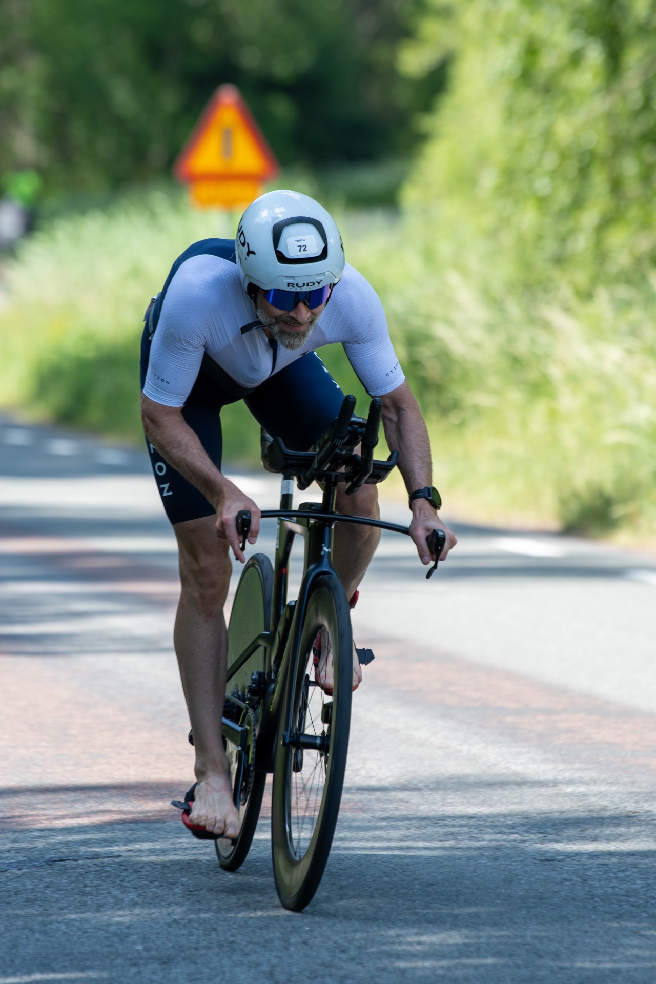 Triathlet på landsväg, på väg till transition, hjälm med nummerlapp, suddig varningsskylt bakom.